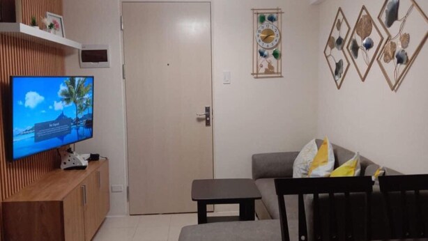 A living room with a television on a wooden cabinet, a sofa with cushions, a small table, and a door. - FULLY FURNISHED 2 BEDROOM CONDO UNIT (Pasig)