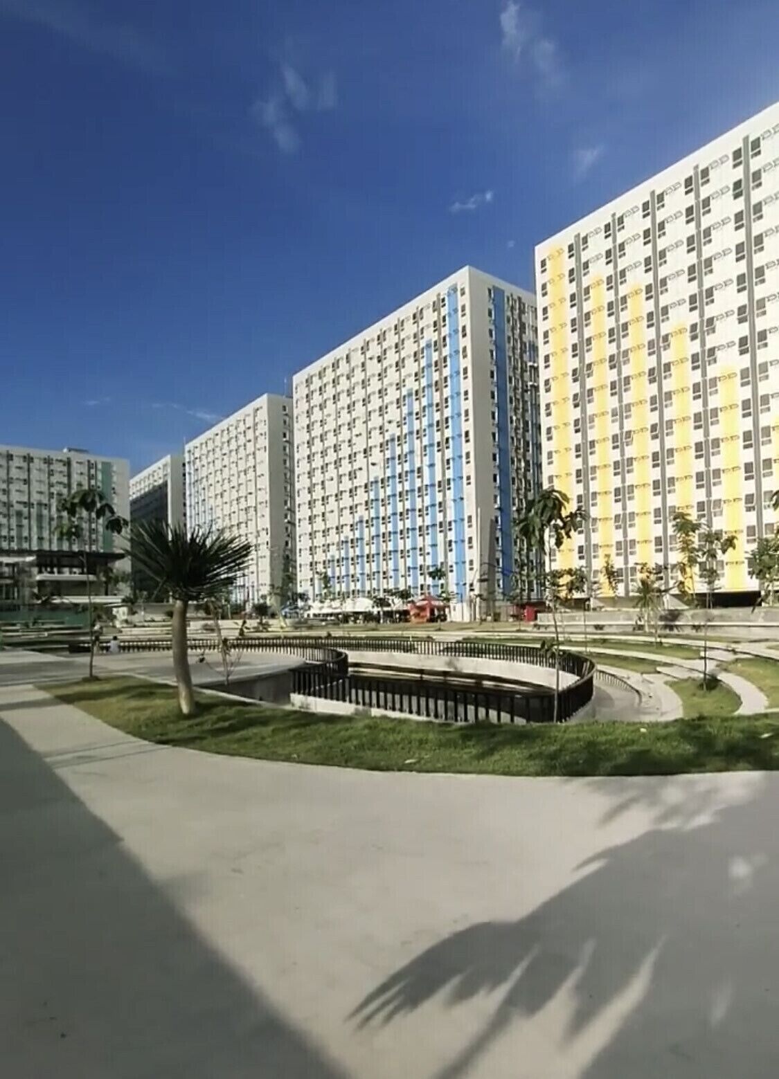 Un immeuble résidentiel moderne de grande hauteur, doté d’une façade uniforme, entouré d’un espace paysager agrémenté de palmiers et d’un bassin circulaire.