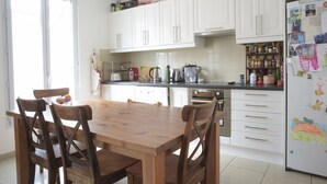 A kitchen with white cabinets, a wooden dining table, and chairs. There are various kitchen appliances and items on the countertops. - Charming town house very close to Paris. (Pantin)
