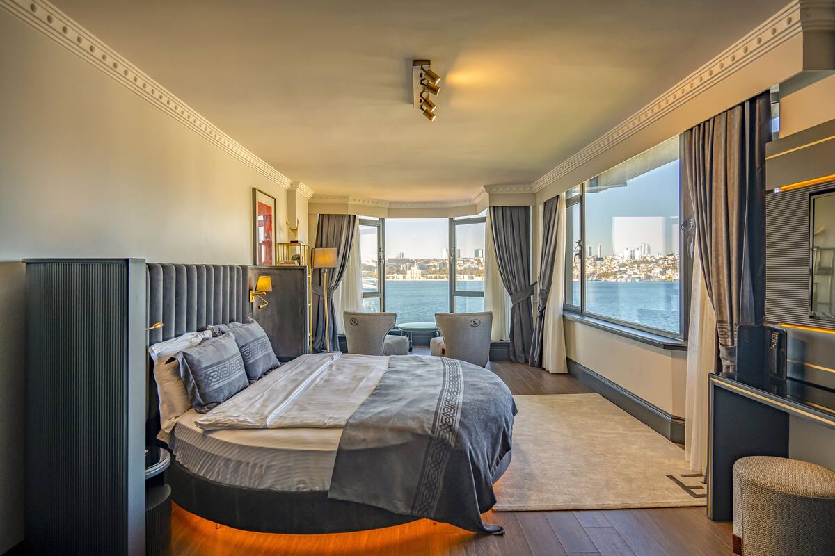 A modern hotel room with a large bed, a seating area, and a view of the city through large windows.