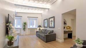 Ein modernes Wohnzimmer mit grauem Sofa, einem Couchtisch und einem Fernseher auf einem Schrank. Zwei Fenster mit Jalousien, eine Decke mit Holzbalken und ein kleiner Schreibtisch mit Lampe sind ebenfalls zu sehen.