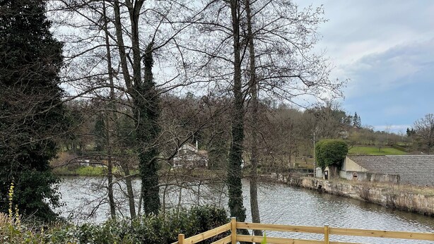 A wooden deck with a picnic table set for four, surrounded by a wooden fence, overlooking a body of water and trees. - Charming house in the forest of Troncais (Saint-Bonnet-Tronçais)