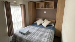 A neatly made bed with a plaid bedspread, two pillows with a leaf pattern, and a folded towel on the bed. There is a wooden shelf above the bed with two small bottles. - Caravan - Sleeps 10 - Pet Friendly - Parking (Clacton-on-Sea)