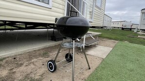 A portable barbecue grill with wheels, set up on a patch of grass next to a mobile home. - Caravan - Sleeps 10 - Pet Friendly - Parking (Clacton-on-Sea)