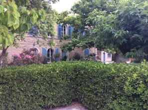 A stone building with arched windows and blue shutters, surrounded by lush greenery and flowering plants.