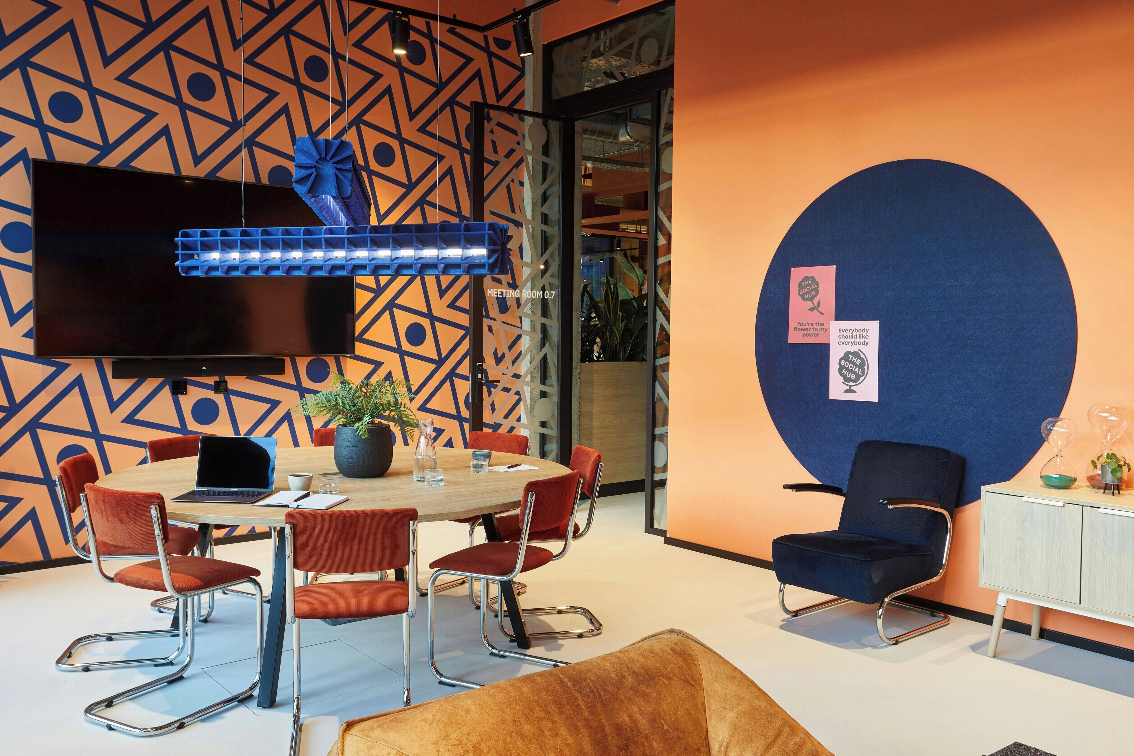 A modern meeting room with a large table, red chairs, a TV, and a decorative wall.