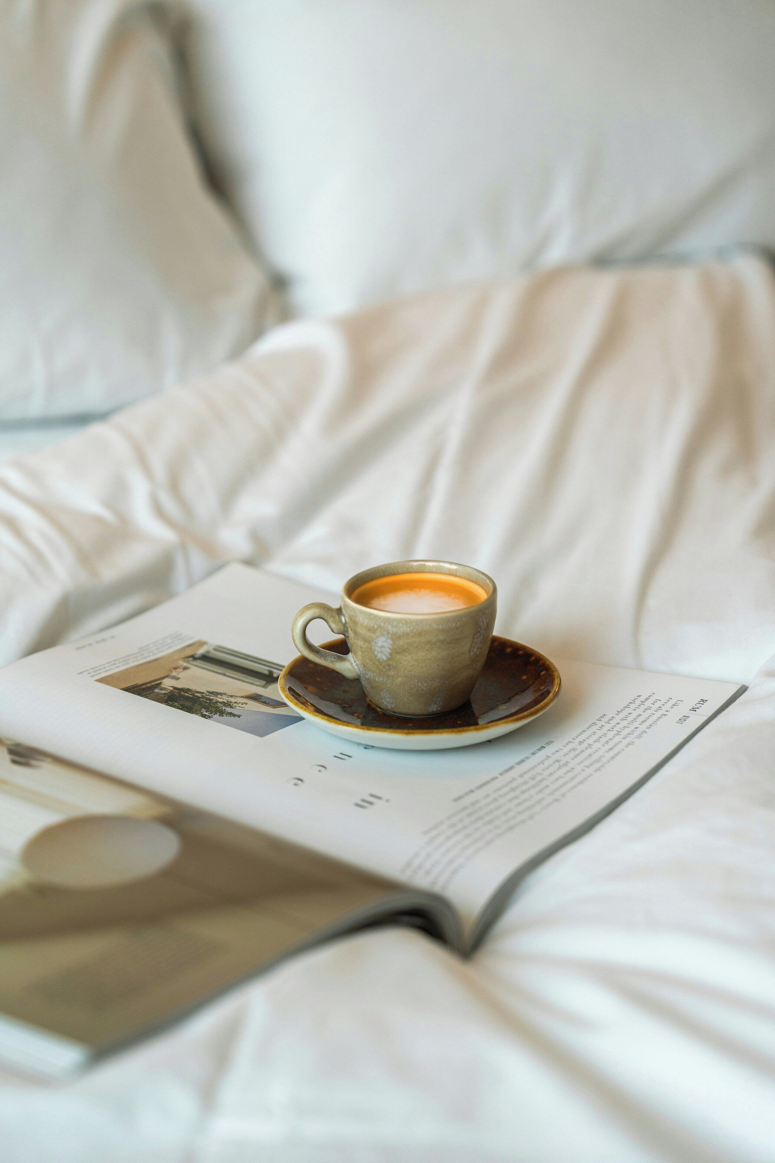 Uma chávena de café sobre um pires, numa cama com uma revista.