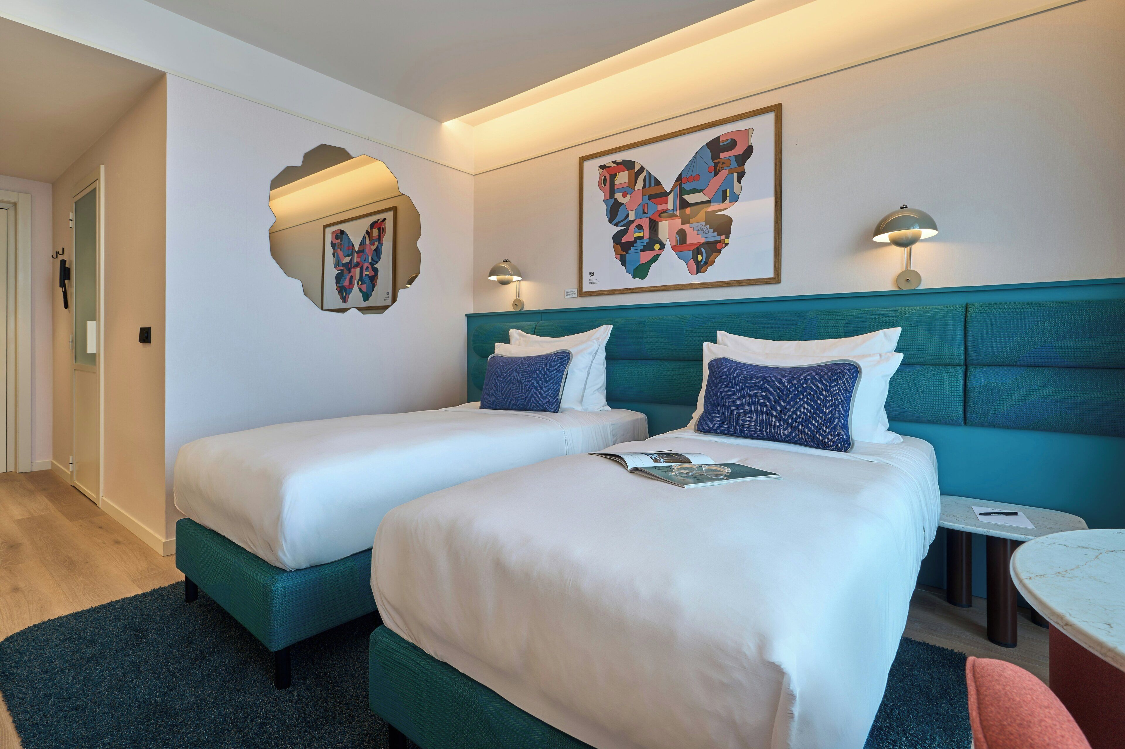 A hotel room with two beds, a teal headboard, a small table, and a wall-mounted lamp.