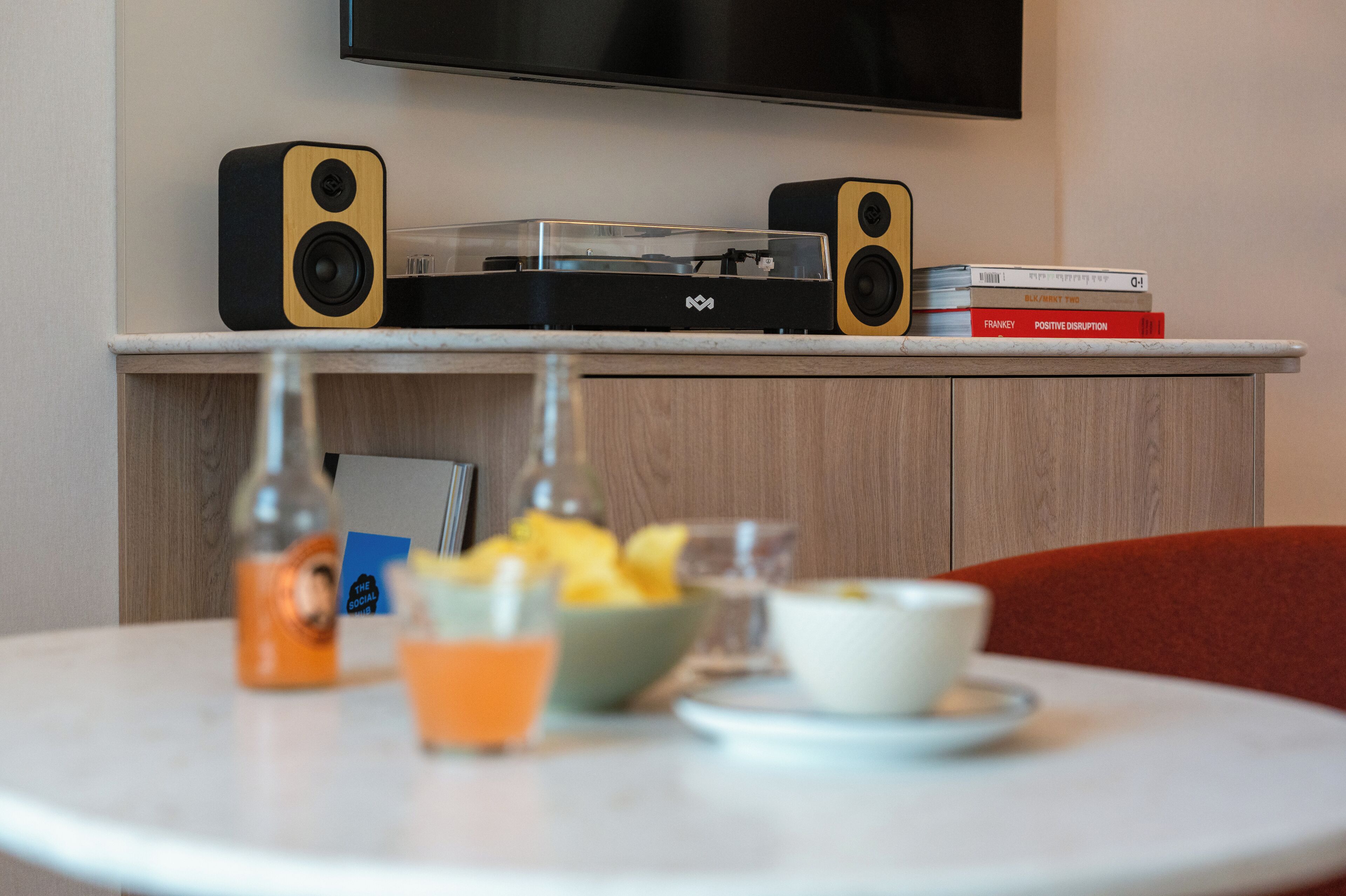 Una habitación con una televisión de pantalla plana montada en la pared, un tocadiscos con altavoces, y una mesa con un frutero, un vaso de zumo de naranja y una botella.