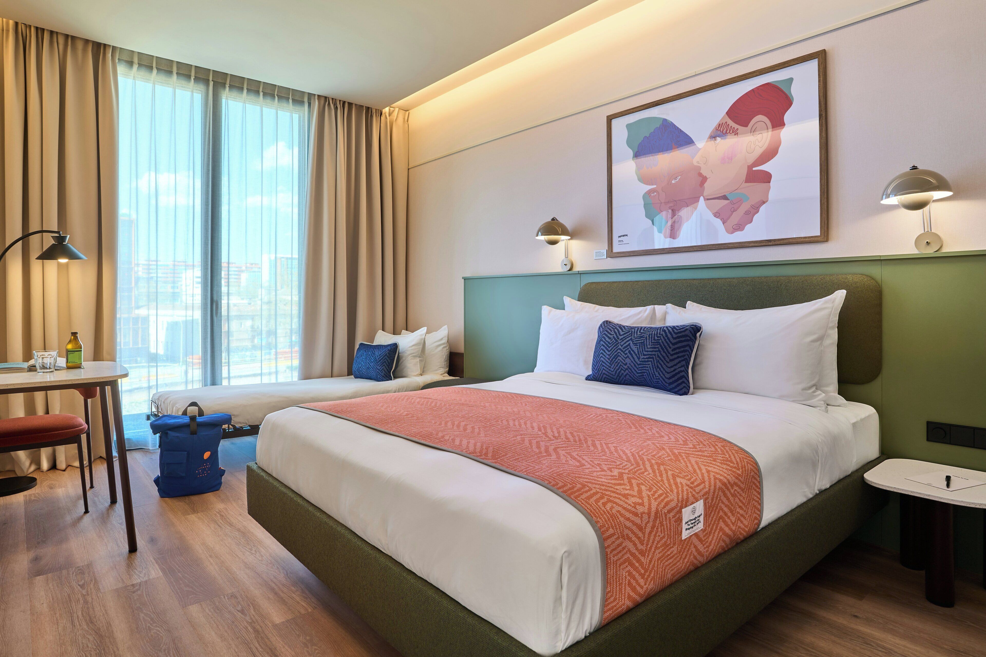 A hotel room with a large bed, a desk with a chair, a window with curtains, and a wall-mounted lamp.