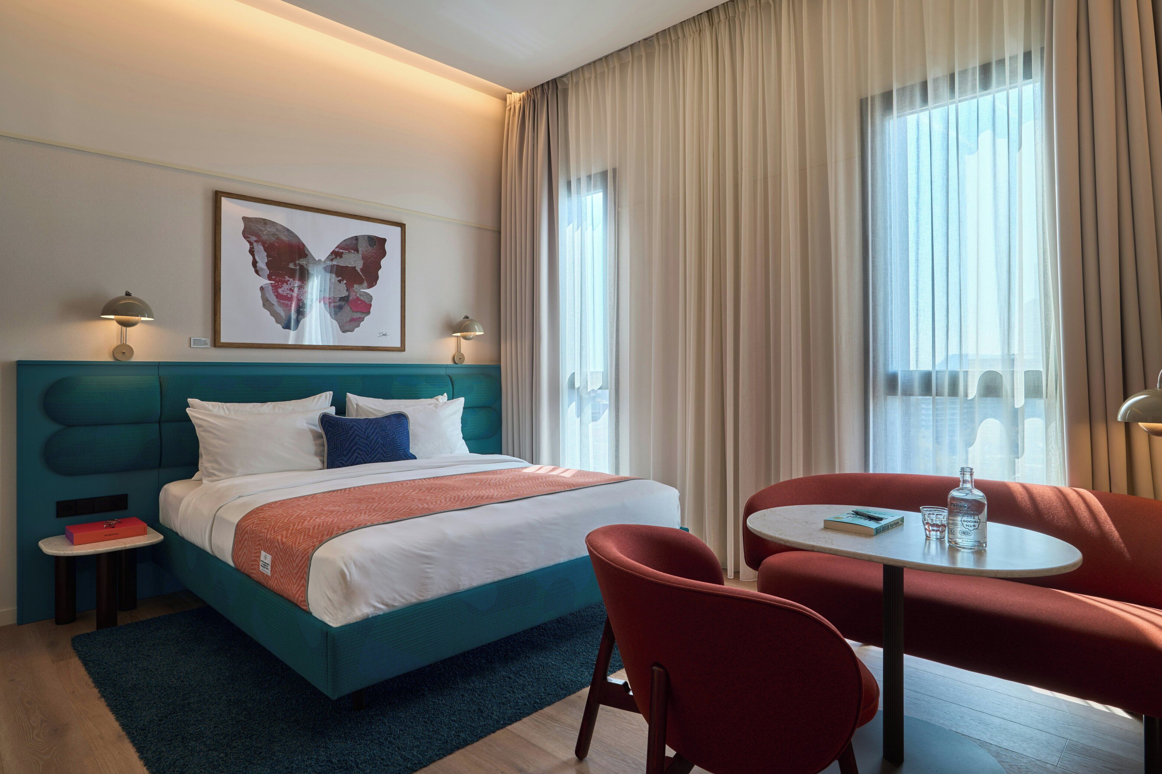 Habitació d'hotel amb un llit, una taula amb cadires, una petita taula auxiliar i una finestra amb cortines.