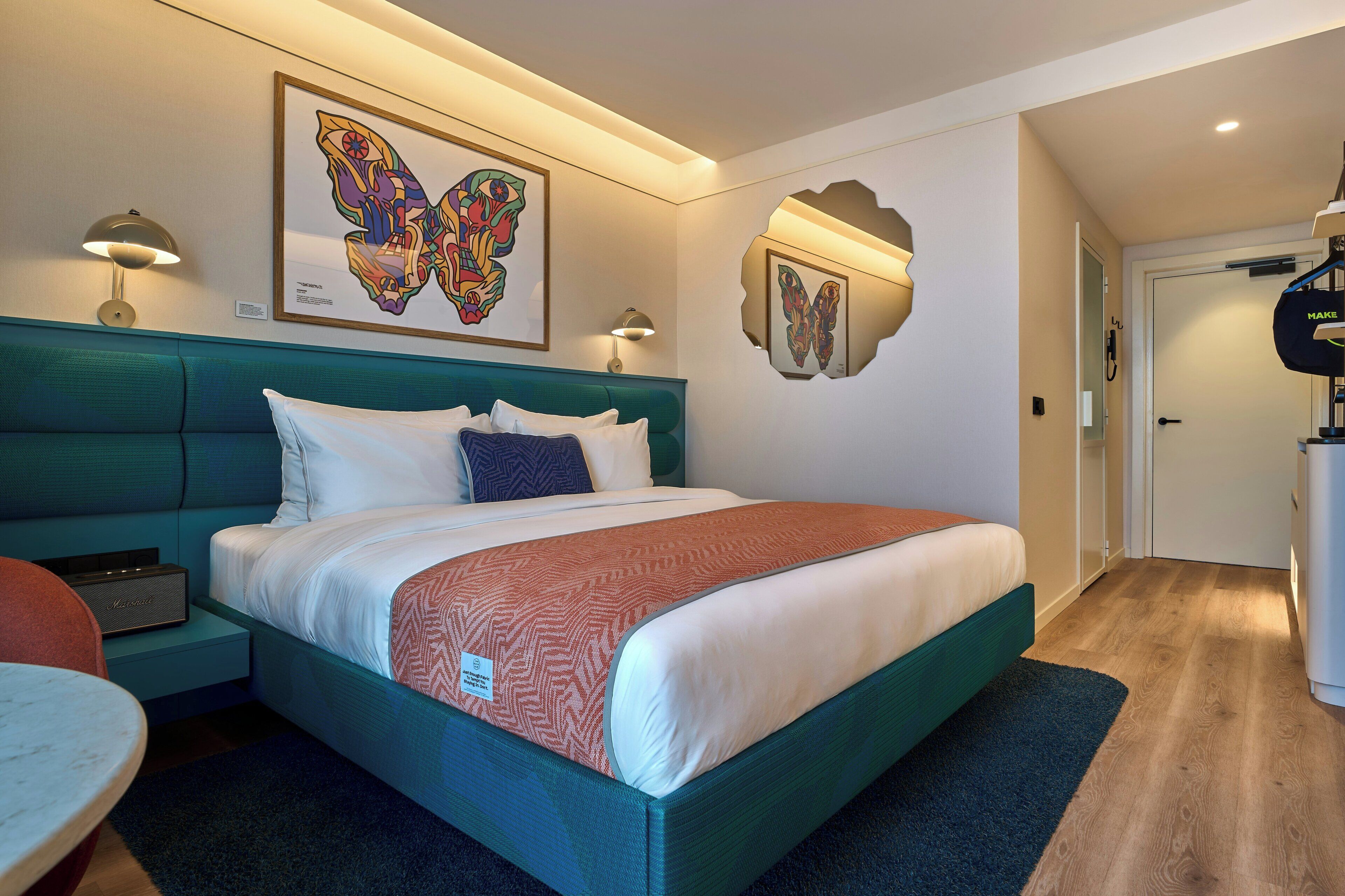 Habitació d'hotel moderna amb un llit color turquesa, roba de llit blanca, un quadre decoratiu d'una papallona i una petita taula auxiliar amb una làmpada.