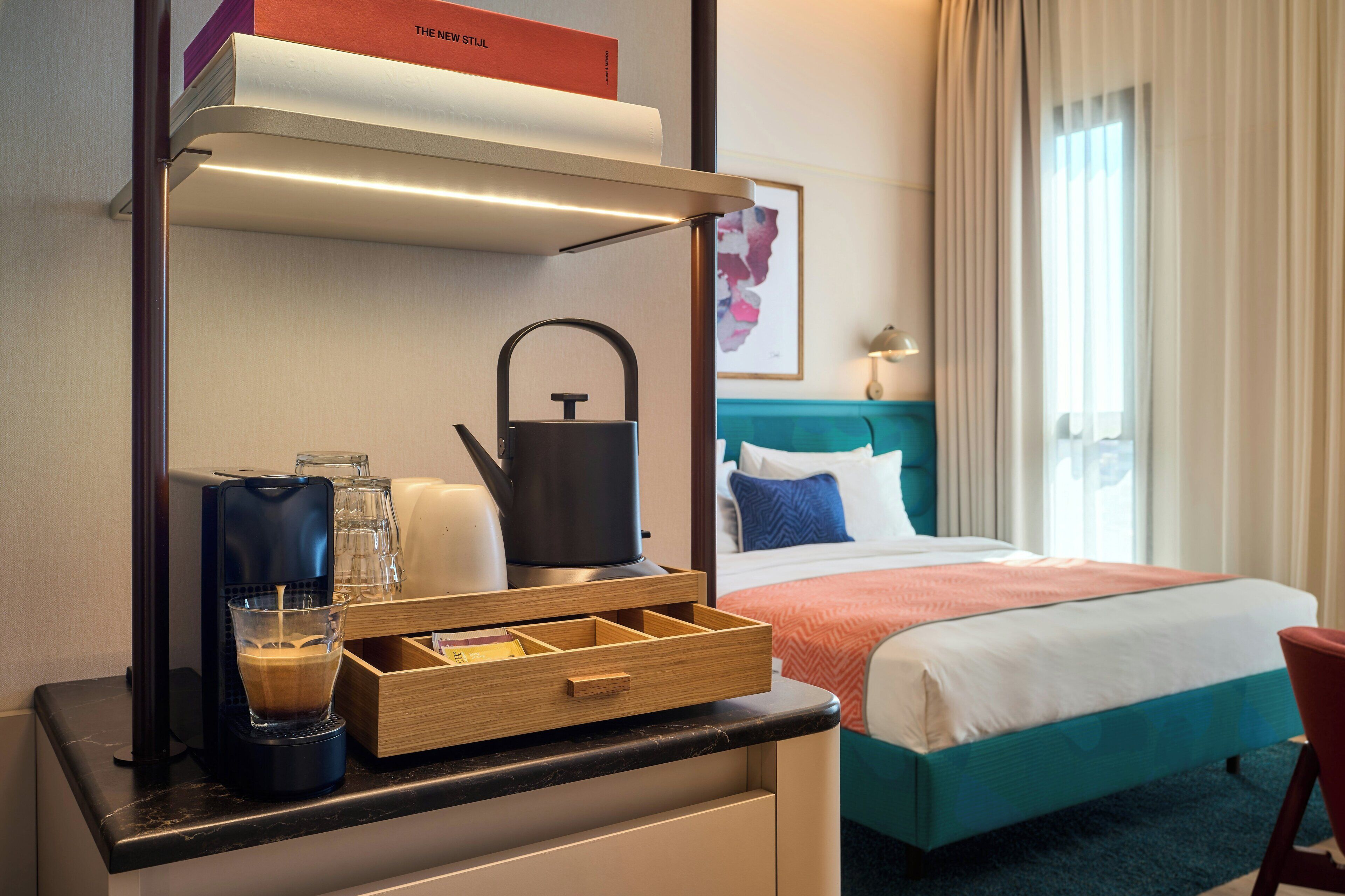 A hotel room with a bed, a desk with a coffee maker, a kettle, and a glass of water.