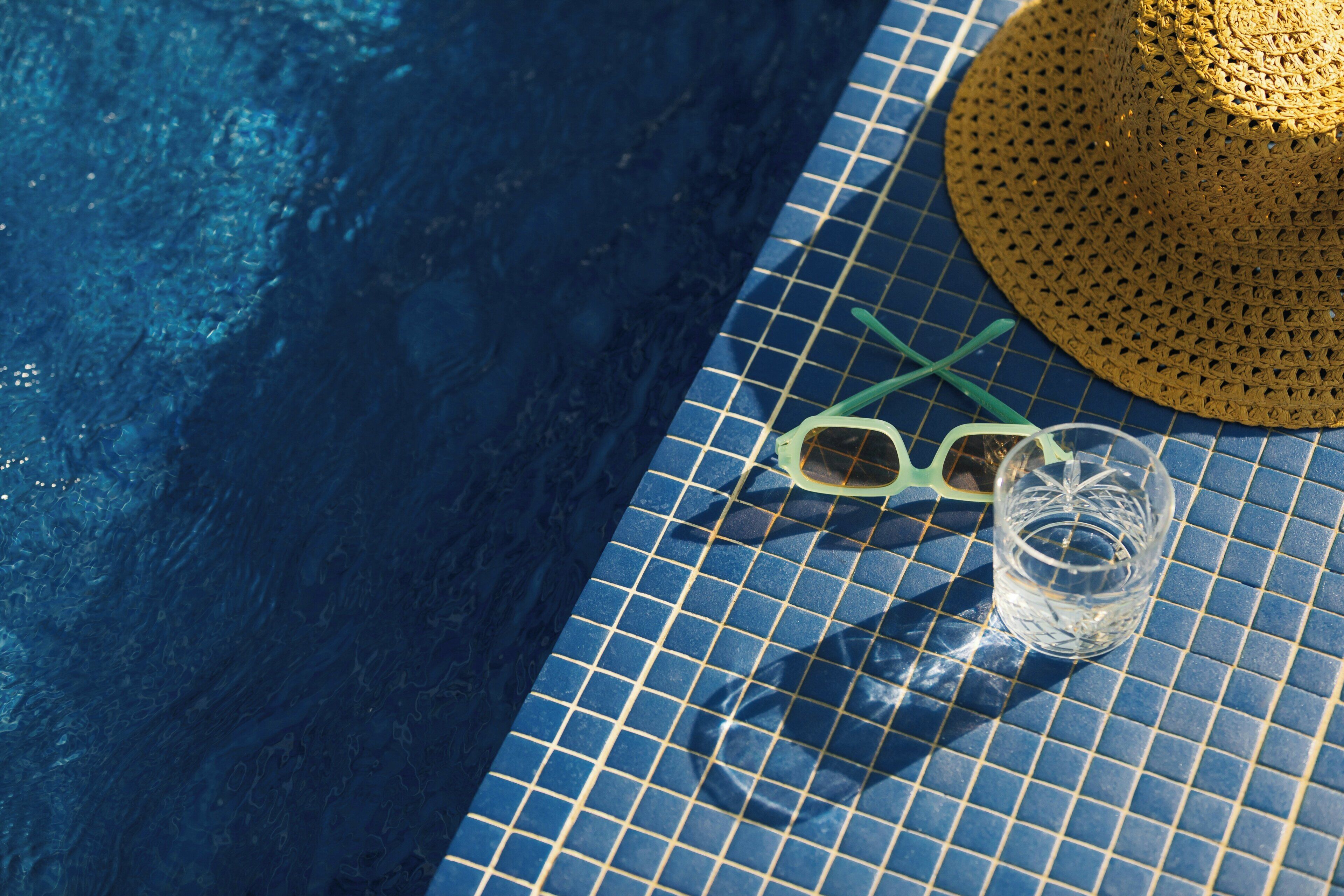 Un área junto a la piscina con una mesa de cerámica, un sombrero de paja, gafas de sol y un vaso de agua.