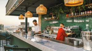 Um bar no terraço com balcão, cadeiras de vime, luminárias pendentes e vista para a cidade.