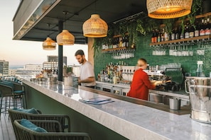A rooftop bar with a bar counter, wicker chairs, hanging wicker lamps, and a view of the cityscape.