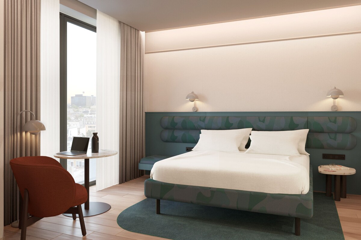 A hotel room with a bed, a chair, a small table, and a view of the city.