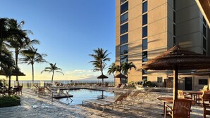 Ein Poolbereich mit Liegestühlen, einem Pool, Palmen und einem Gebäude mit Blick aufs Meer.