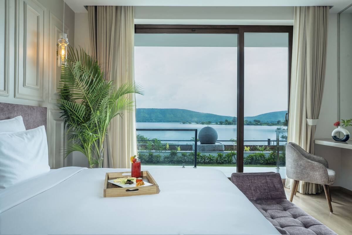 Une chambre d’hôtel avec un lit, un plateau avec de la nourriture et des boissons, une chaise, une grande fenêtre donnant sur un lac et des montagnes.