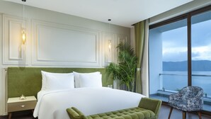 A hotel room with a large bed, a green bench, a chair, and a view of the sea through large windows. - Zana Lake Resort Udaipur (Udaipur)