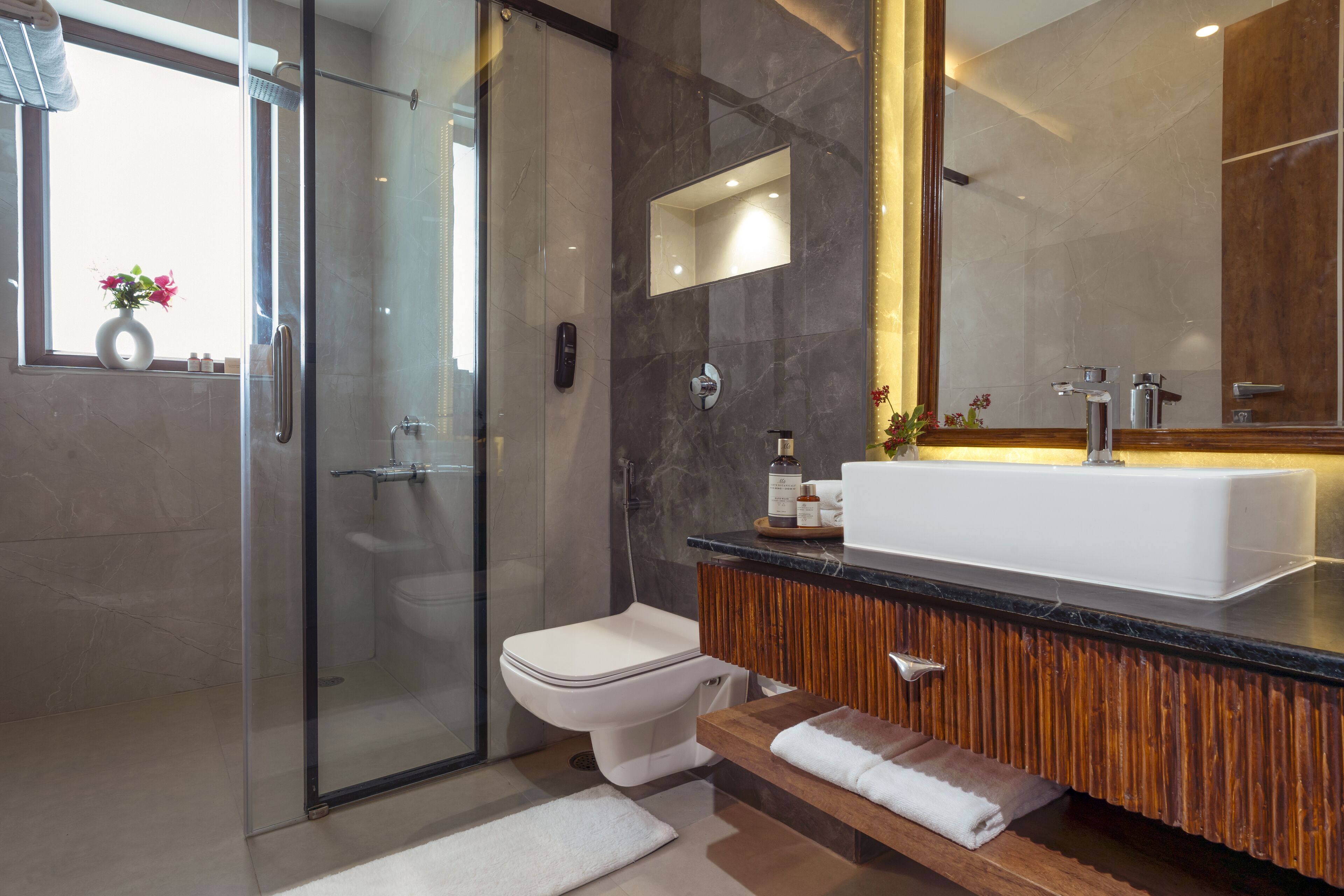A modern bathroom with a glass shower enclosure, a white toilet, a wooden vanity with a large mirror, and a white sink.