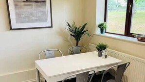 A dining area with a white table and four chairs, a framed picture on the wall, a window with a view of greenery, and a radiator below. - Cosy Wrekin View Annexe (Eaton Constantine)