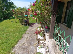Un sendero de piedra que lleva a una casa con baranda verde, un pequeño jardín con flores y una estructura de techo de paja.