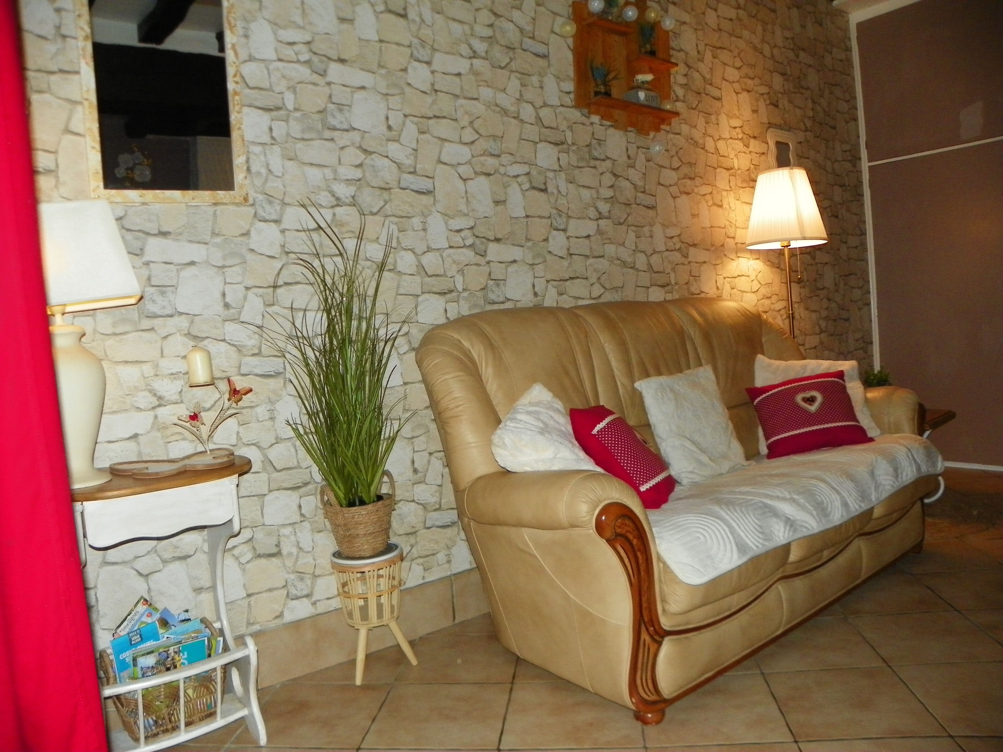 Ein Wohnzimmer mit einer Steinsäule, einem beige farbenen Sofa mit Kissen, einem kleinen Beistelltisch mit Lampe und einer Topfpflanze.