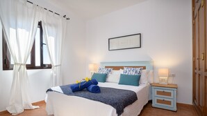 A neatly made bed with a white and blue bedspread, two pillows, and a blue blanket with yellow flowers. There is a small bedside table with a lamp and a wall-mounted picture above the bed.