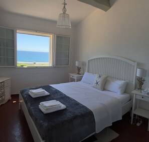 A bedroom with a bed, two bedside tables, a nightstand, a window with a view of the sea, and a hanging light fixture.