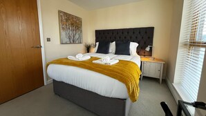 A neatly made bed with a yellow throw blanket and two towels, a dark headboard, a bedside table with a lamp, and a window with blinds.
