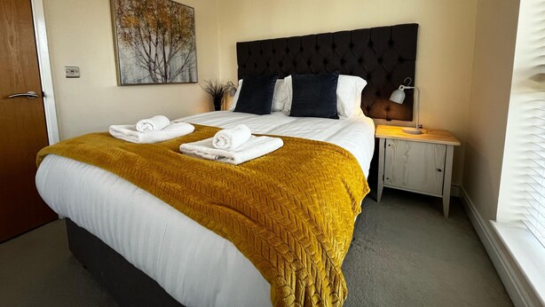 A hotel room with a large bed, a headboard, a nightstand, a lamp, a painting, and a window with blinds.