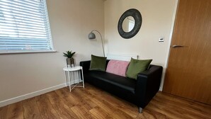 A living room with a black sofa, a small white side table, a potted plant, a floor lamp, a round mirror, and a wooden door.