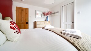 A neatly made bed with white linens and a red embroidered pillow, a wooden dresser with a mirror above it, and a white door with a handle. - A beautiful renovated Georgian house minutes walk to Port Isaac's harbourside (Port Isaac)