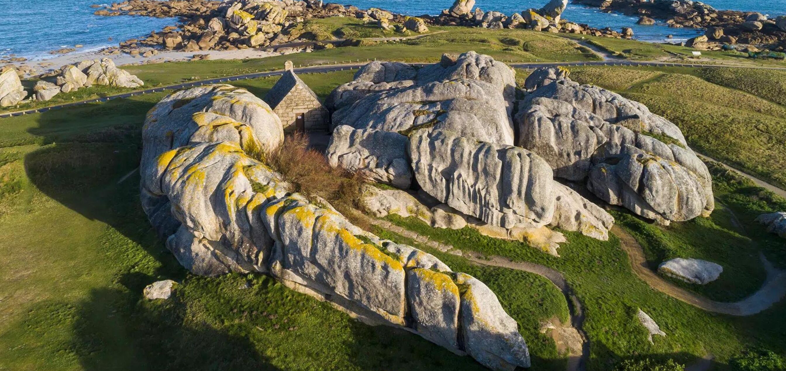 Une grande formation rocheuse abrite un petit bâtiment en pierre au centre, le tout étant entouré d’une pelouse verdoyante et d’un chemin pavé.