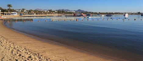 Ein Strand mit klarem, blauem Wasser, einem sandigen Ufer und Booten in der Ferne.
