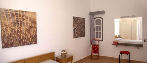 A bedroom with a bed, a small table with a decorative item, a window with shutters, and a wall-mounted shelf.