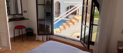 Un dormitorio con una cama grande, una silla roja, una mesita, un espejo y una ventana con vistas a una piscina y unas escaleras.