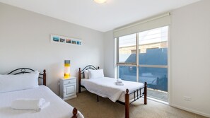 Two single beds with white bedding and matching nightstands, each with a lamp, are situated near a large window offering a view of the outside.