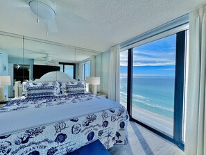 Quarto com cama grande, ventilador de teto, mesa de cabeceira, luminária e vista para o oceano.