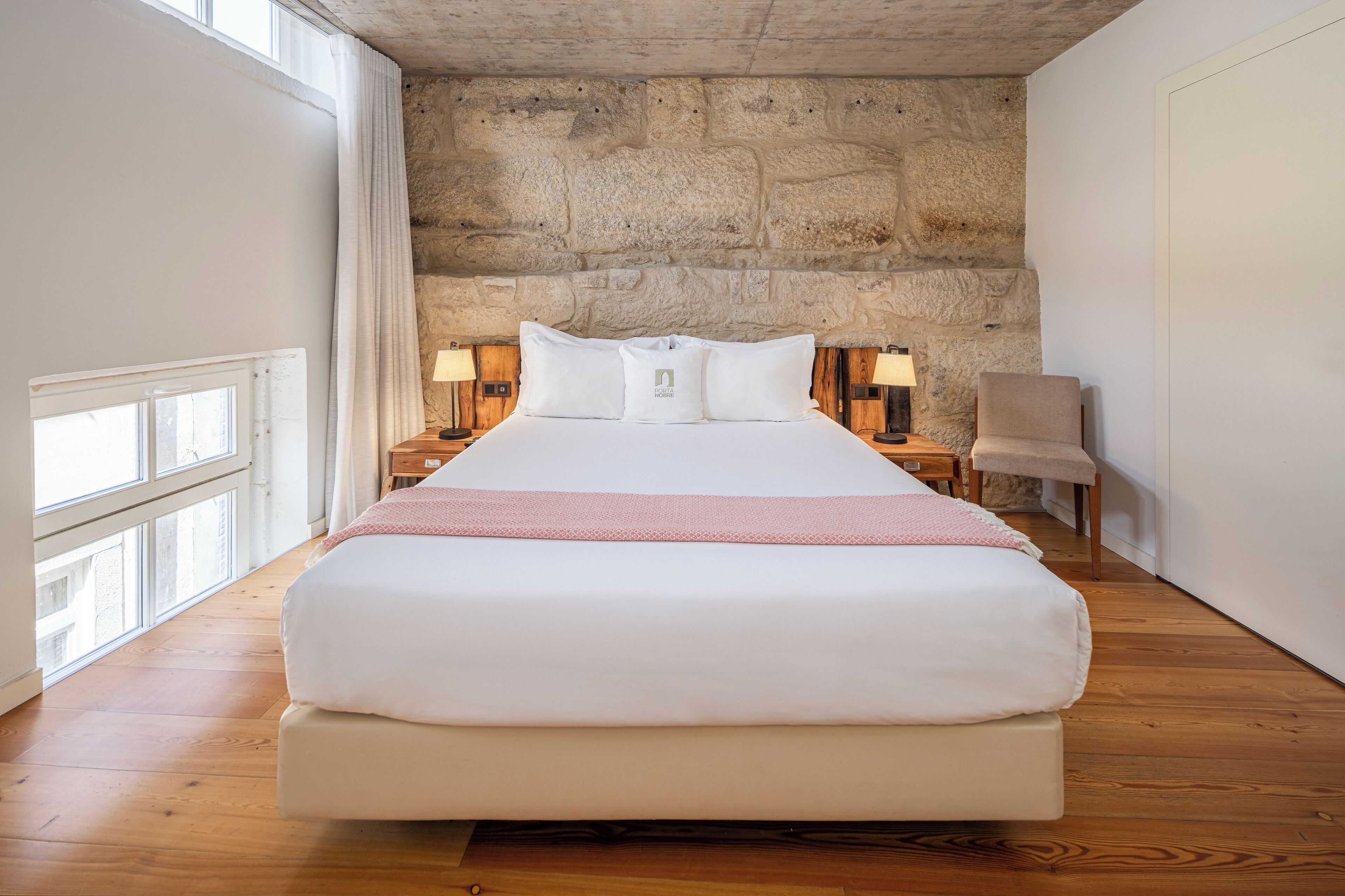 Una cama grande con sábanas blancas, un muro de piedra al fondo y dos lámparas de noche.