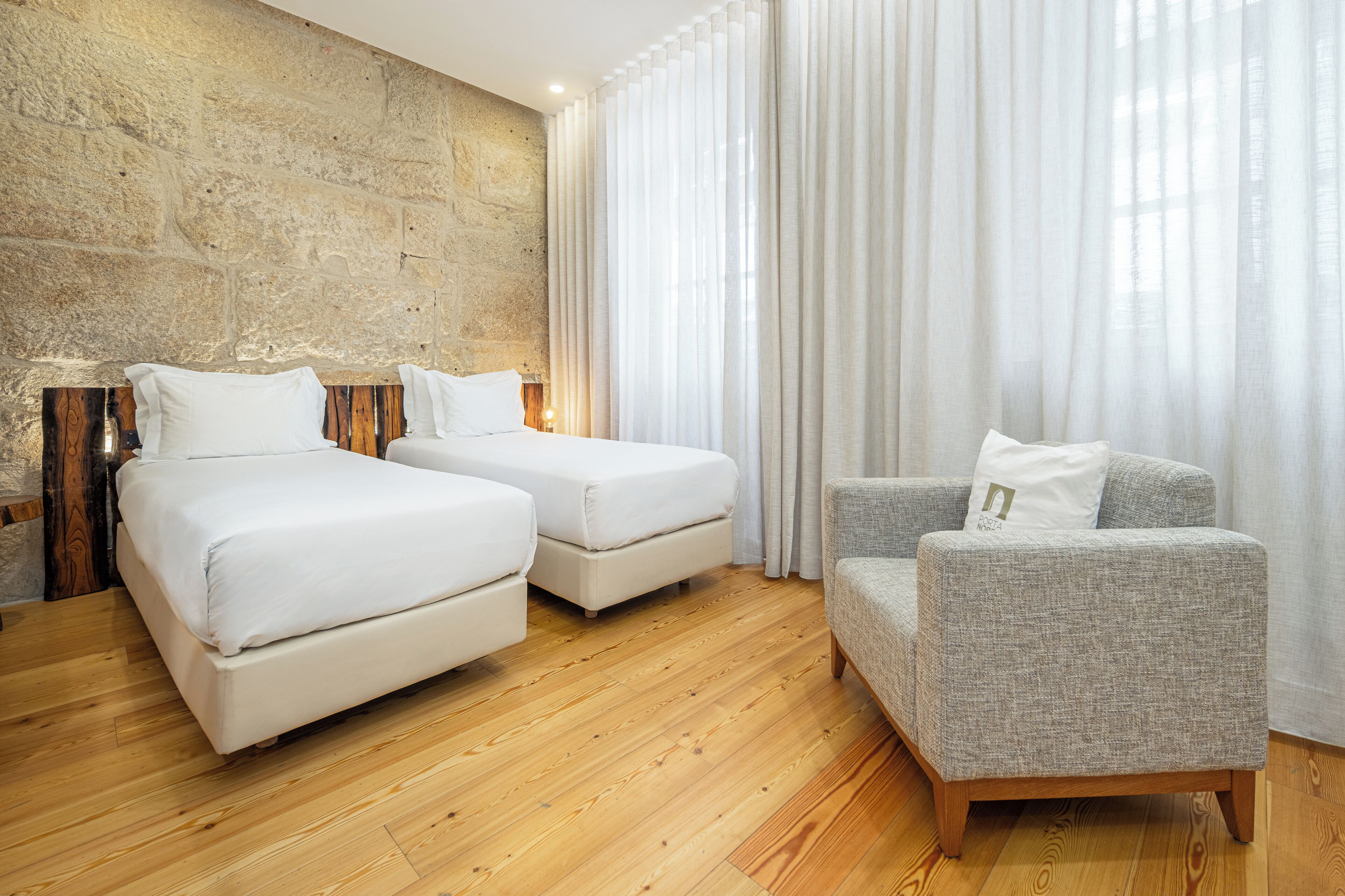 Ein Zimmer mit zwei Betten, einem grauen Sessel, Holzböden und einer Steinwand.
