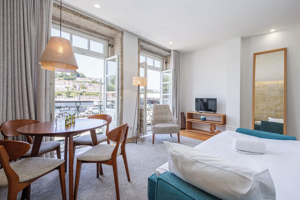 A hotel room with a bed, a TV, a dining table with chairs, and a view of the city.