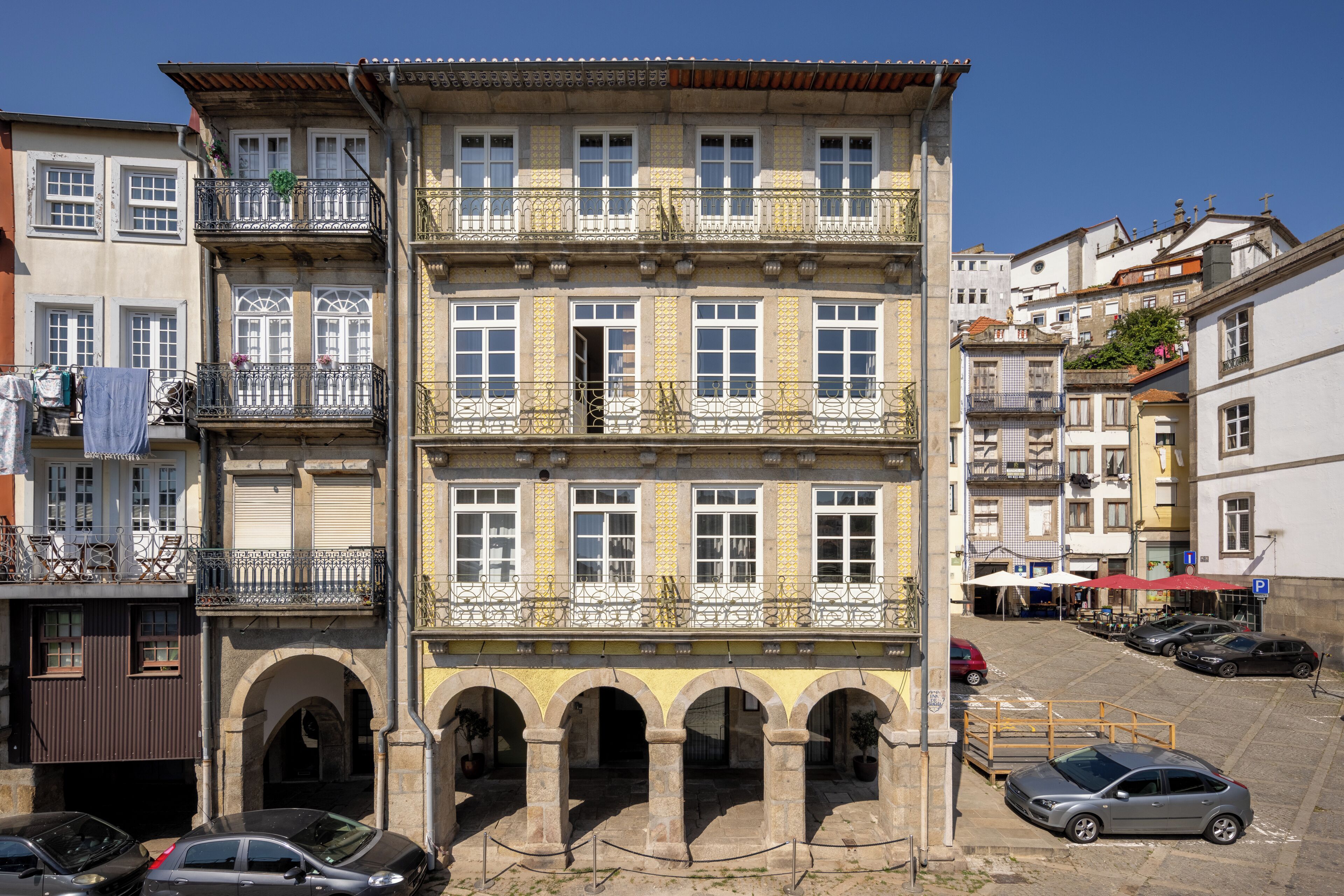 A multi-story building with balconies, arched entrances, and a parking area with cars.