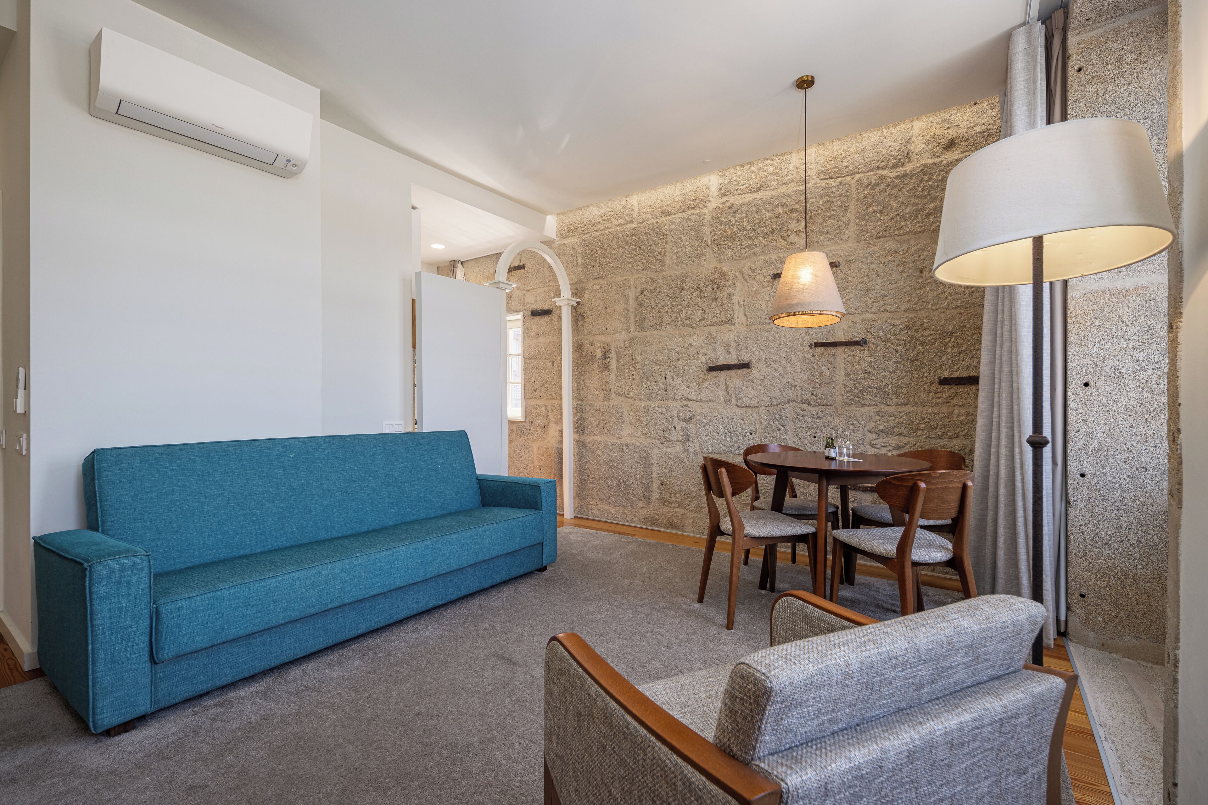 Quarto com sofá azul, mesa de jantar com cadeiras, luminária e parede de pedra.