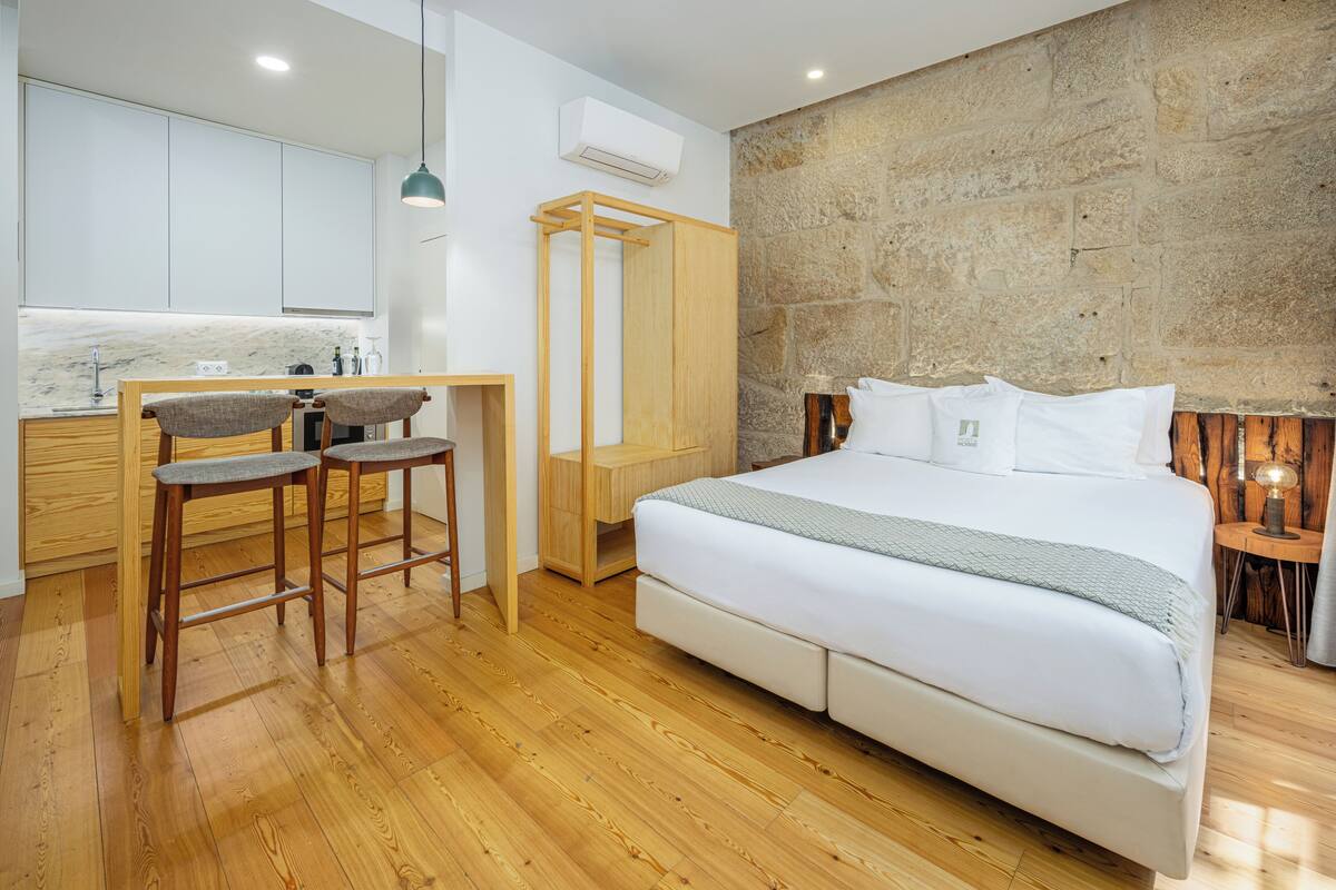 A modern hotel room with a wooden floor, a bed with white bedding, a small kitchen area with a marble countertop, and a stone accent wall.