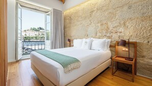 A bedroom with a large bed, a wooden nightstand, a stone wall, and a view of buildings through an open door. - Porta Nobre Boutique Hotel by Aspasios (Porto)