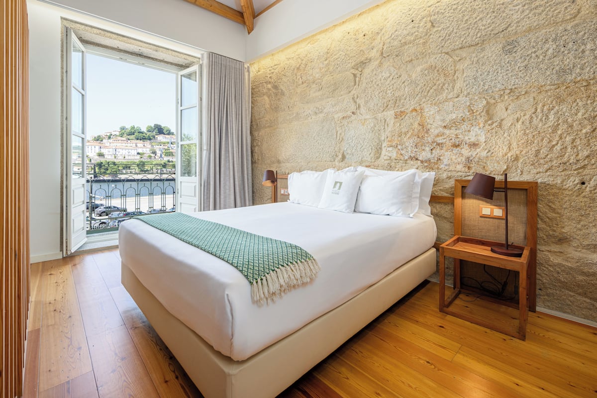 A bedroom with a large bed, a wooden nightstand, a stone wall, and a view of buildings through an open door.