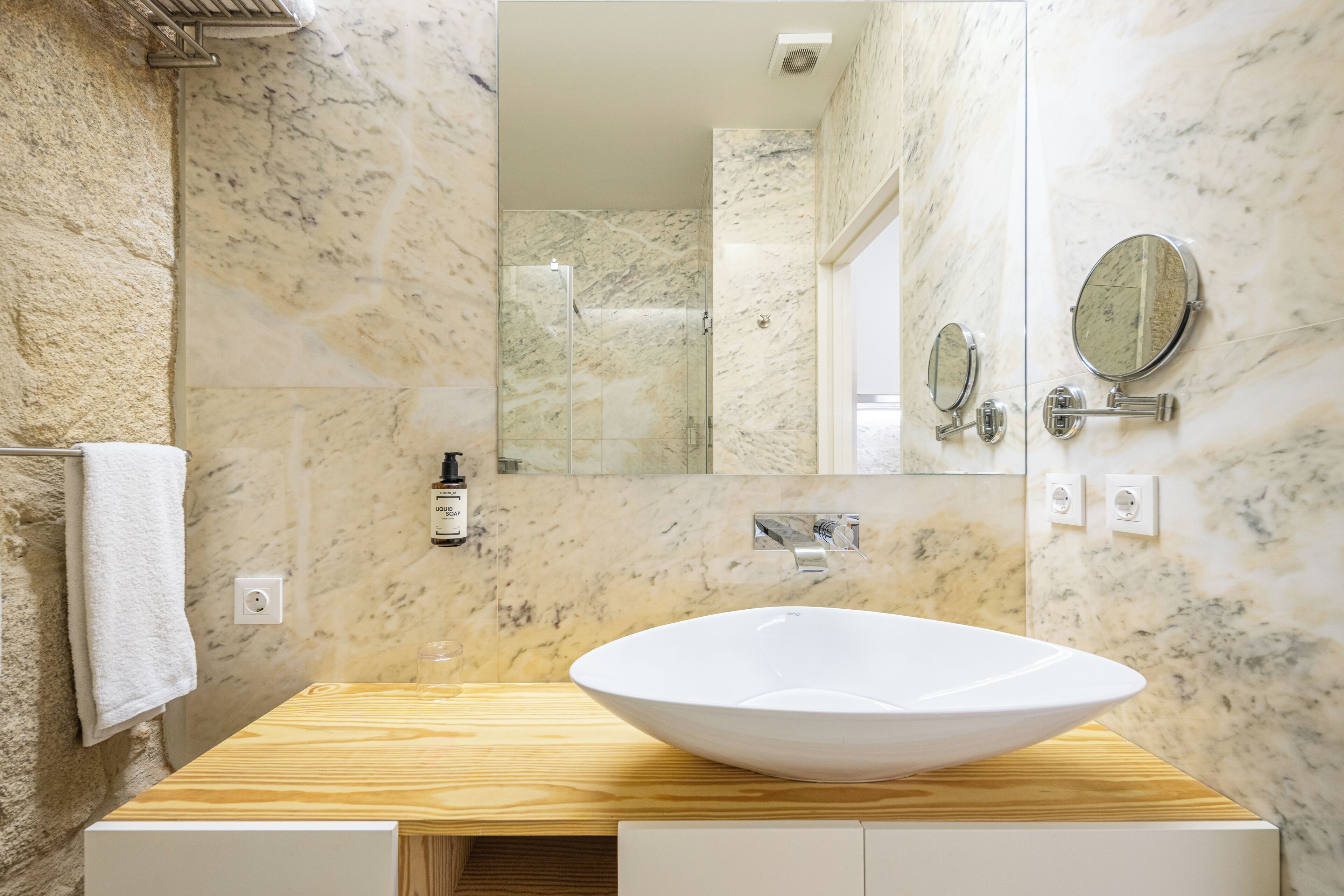 A bathroom with a white sink, marble walls, a glass shower enclosure, and a mirror.