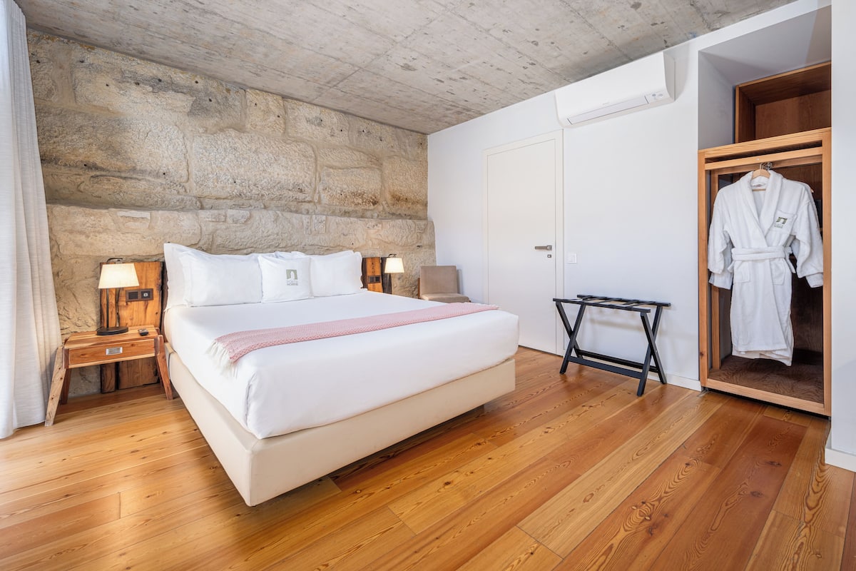 A bedroom with a large bed, wooden flooring, a stone wall, a wooden wardrobe, and a robe hanging on the door.